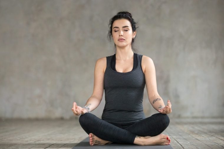 Young sporty woman practicing yoga, doing Sukhasana exercise, Easy Seat pose, working out, wearing sportswear, black pants and top, indoor full length, yoga studio