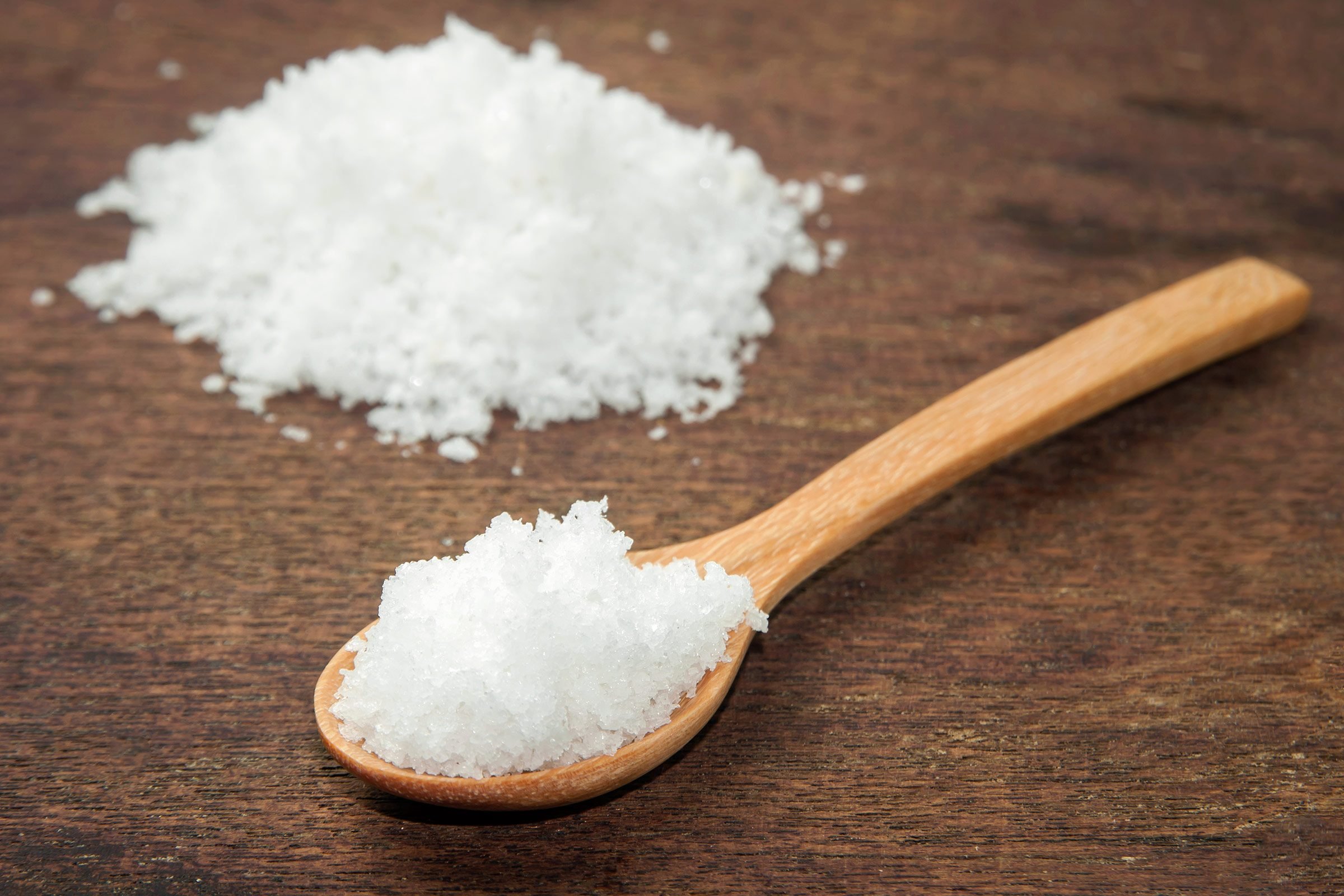 Wooden spoon of salt on a wooden table. Pile of salt behind it.