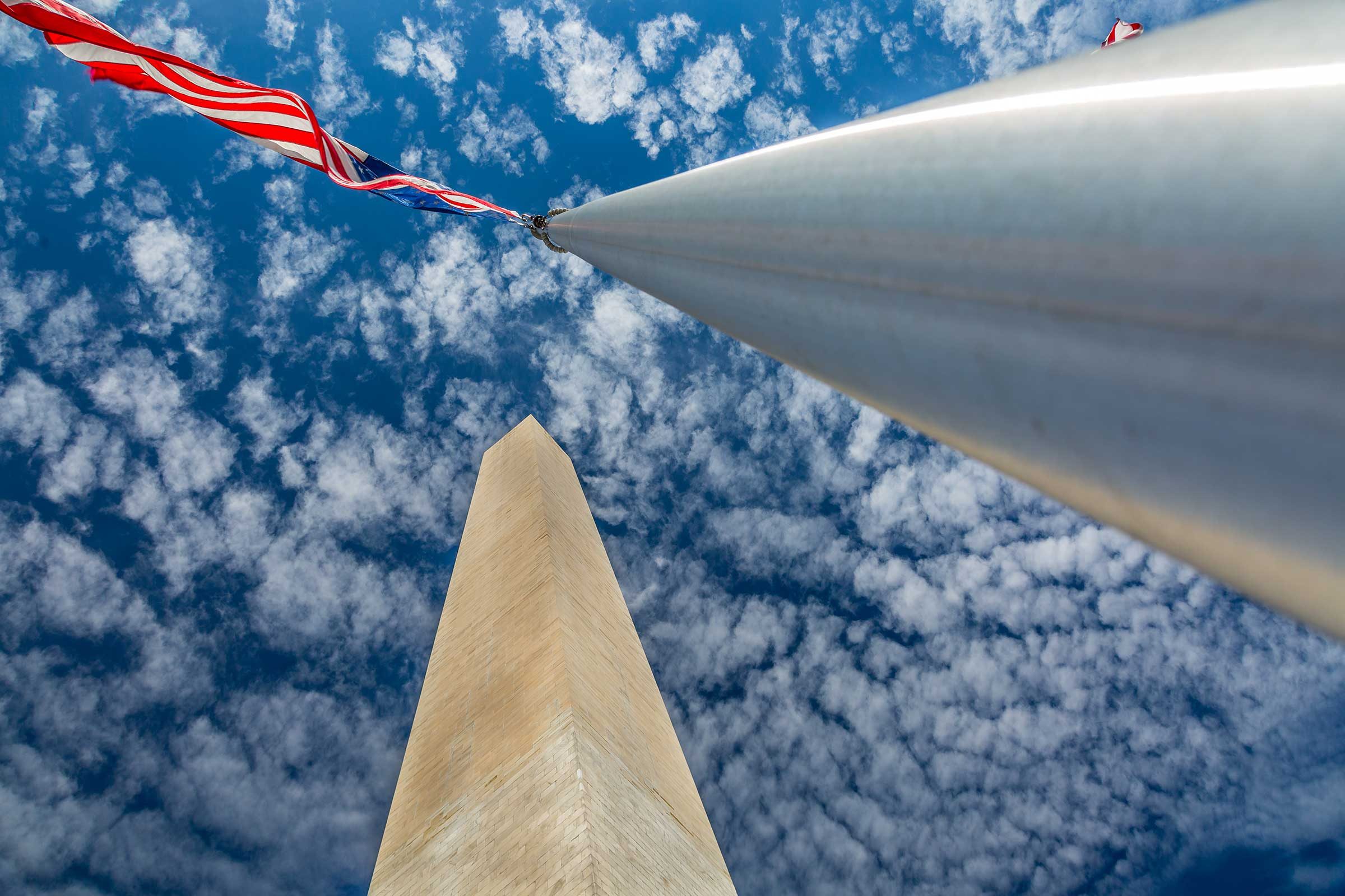 the washington monument in washington, dc, as seen from the base of one of the flag poles. an american flag waves below a blue sky dotted with fluffly clouds