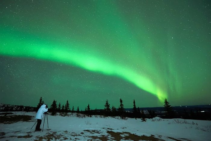 a person in a snowy landscape with a camera on a tripod viewing the green skies from the northern lights
