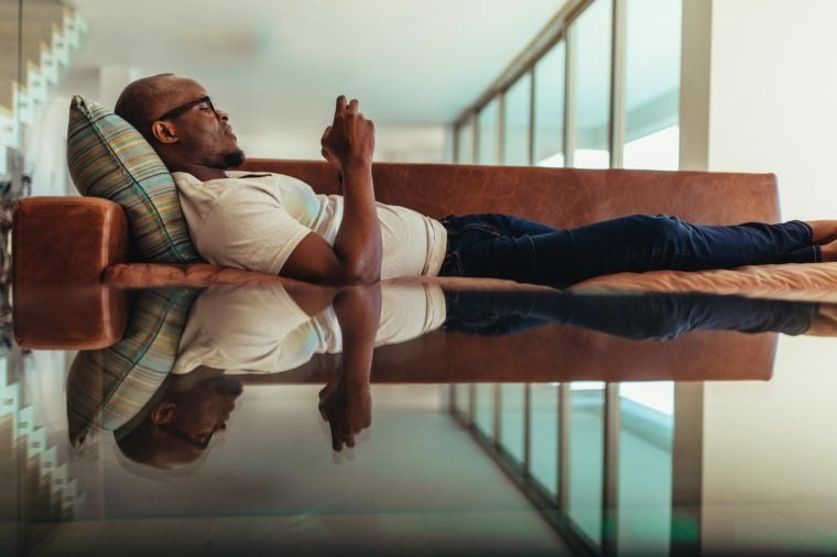 Man relaxing on a lounge and operating mobile phone. Symmetrically opposite reflection of a man relaxing on lounge falling on glass top.