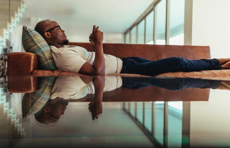 Man relaxing on a lounge and operating mobile phone. Symmetrically opposite reflection of a man relaxing on lounge falling on glass top.