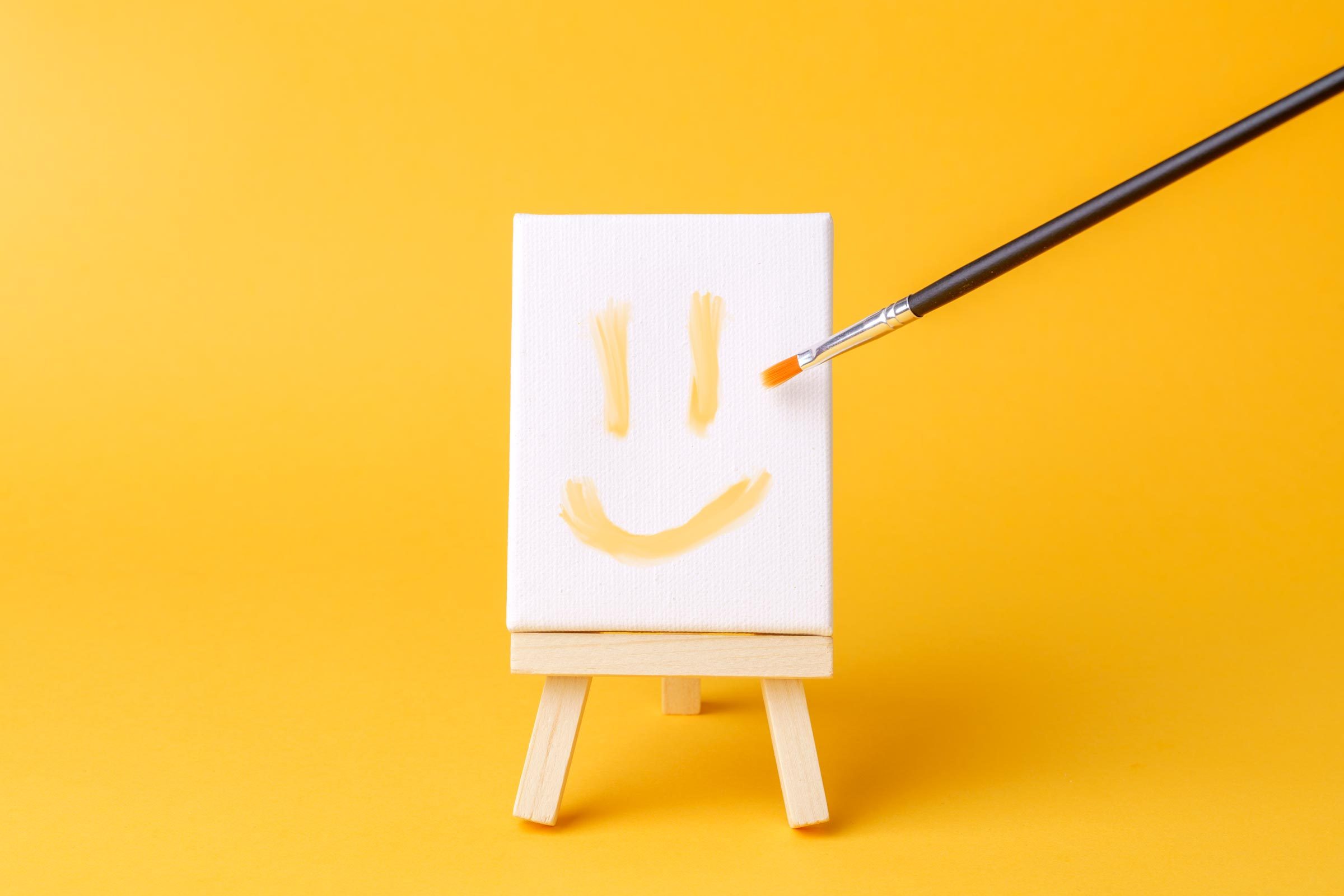 a smiley face painted on a miniature canvas on an aisle with a paint brush on a yellow background