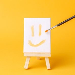 a smiley face painted on a miniature canvas on an aisle with a paint brush on a yellow background