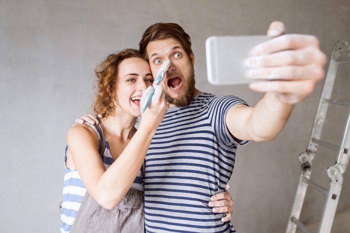 Beautiful young couple in love having fun, painting walls of their new house, taking selfie with smart phone. Home makeover and renovation concept.
