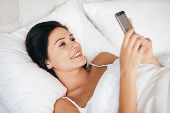 Always available for him. Beautiful young woman smiling and holding smart phone while lying in the bed at home