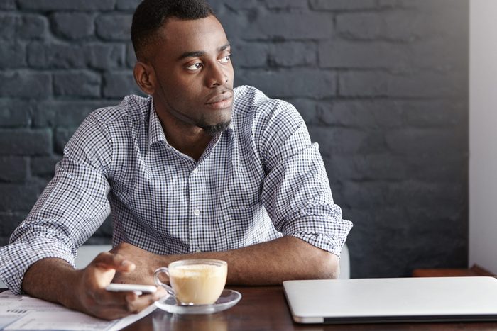 Attractive young African-American entrepreneur typing sms on smart phone with thoughtful and serious look, sitting alone at cafe table with mug, papers and laptop computer, waiting for his lunch