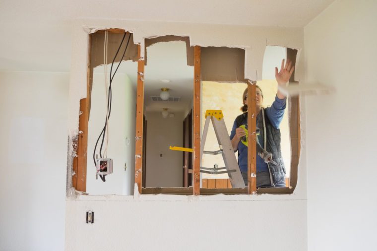 Female construction worker and homeowner using a sawsall tool to cut drywall out and take a wall down to a half-wall during a DIY house renovation.