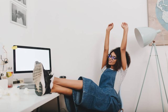 Pretty girl in vintage denim clothes relaxing with legs on table and hands up. Tired young woman in black sneakers resting in office after hard work day.