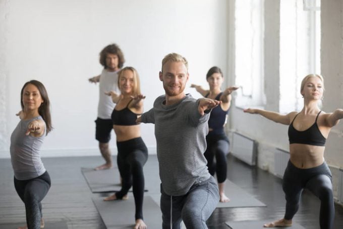 Group of young sporty smiling people practicing yoga lesson with instructor, standing in Warrior Two exercise, Virabhadrasana 2 pose, working out, indoor close up image, studio. Wellbeing concept 