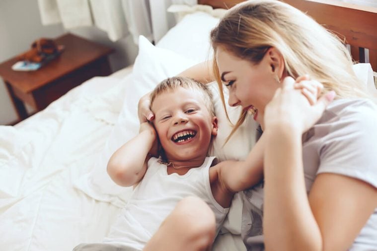 Young mother with her 6 years old little son dressed in pajamas are relaxing and playing in the bed at the weekend together, lazy morning, warm and cozy scene.