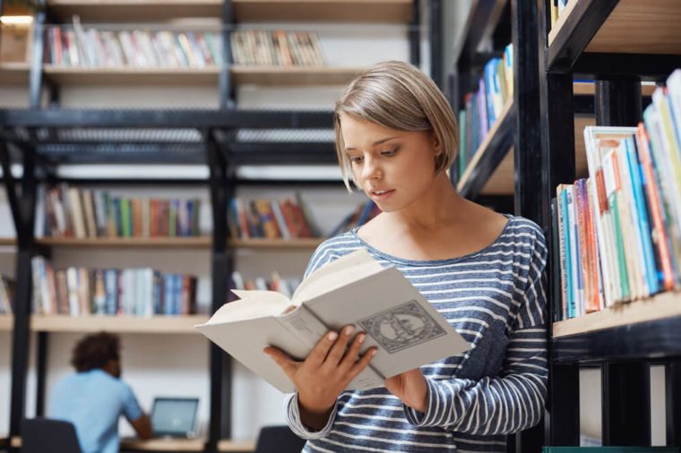 Portrait of charming blonde student girl with short hair in casual clothes standing near shelf in library, reading book, looking through information about economic systems.