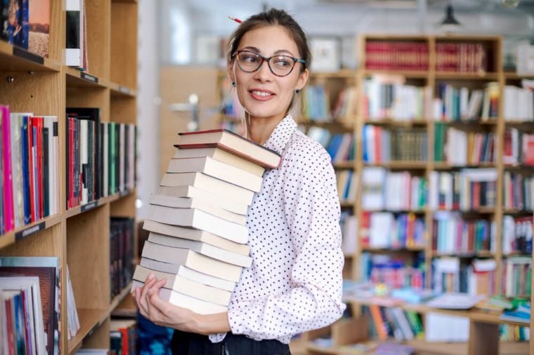 Attractive young woman portrait wearing pink pale shirt with dots and fashionable glasses holding a stack of books in the library surrounded by bookshelves with colorful books.