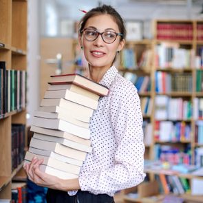 Attractive young woman portrait wearing pink pale shirt with dots and fashionable glasses holding a stack of books in the library surrounded by bookshelves with colorful books.
