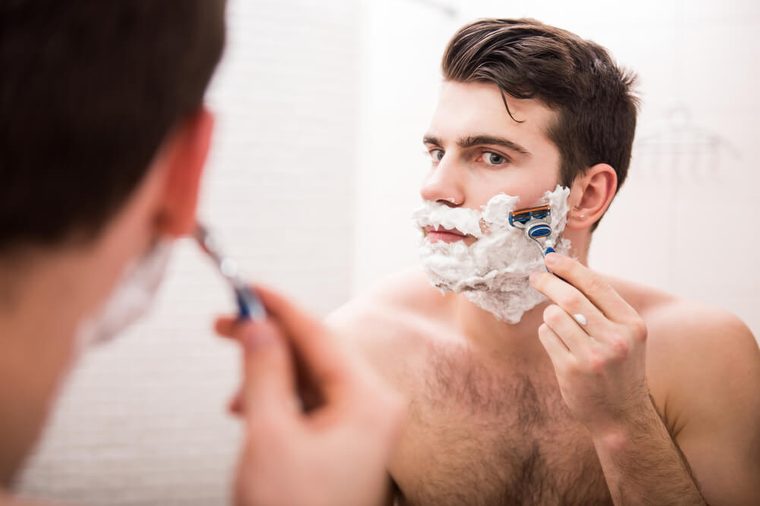 Handsome young man is shaving his face and looking at the mirror.