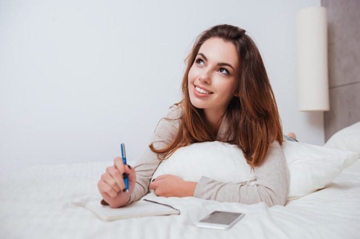 Smart pensive girl lying in bed and making notes in notebook at home