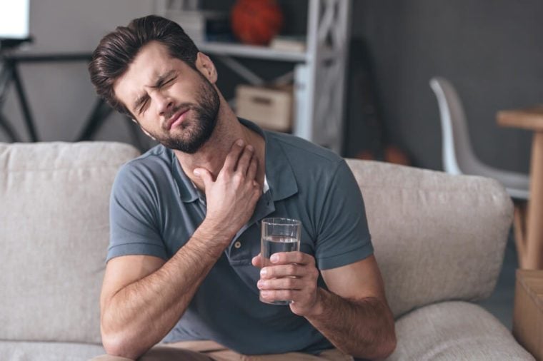 Terrible pain in his throat. Frustrated handsome young man touching his neck and holding a glass of water while sitting on the couch at home