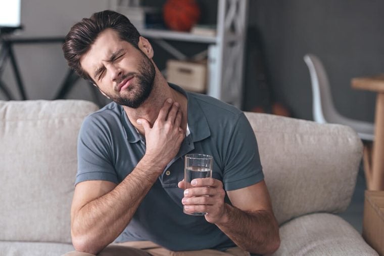 Terrible pain in his throat. Frustrated handsome young man touching his neck and holding a glass of water while sitting on the couch at home