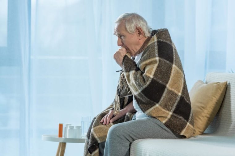 Senior chilled man covered in plaid coughs while sitting sofa in room with medications on table
