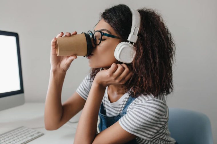 Elegant black girl with curly hairstyle drinking coffee at workplace posing near gray wall. Portrait of tired young woman in white headphones enjoying coffee beside computer.