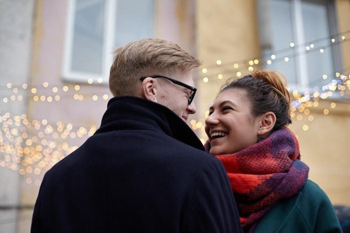 Outdoor portrait of happy smiling couple in love having fun together. Young Caucasian man with tenderness looks at his laughing Latin beloved on the background of garlands of lights