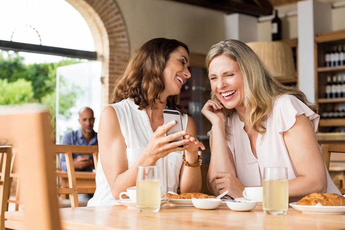Cheerful mature women enjoying a funny video on mobile phone. Mature friends reading a funny message over smartphone. Mid woman showing a cellphone to her friend while laughing over breakfast.