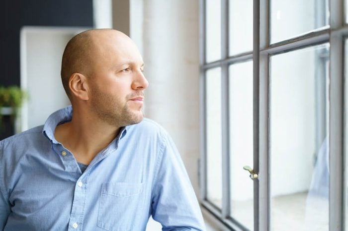Middle-aged man watching through a window with a thoughtful expression as he leans on the sill
