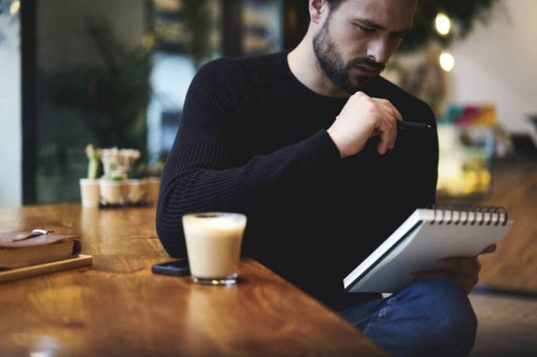 Handsome young designer writes creative graphic notes and making sketch in notebook while sitting at wooden table in coffee interior. Thoughtful man writing new successful ideas for blog article