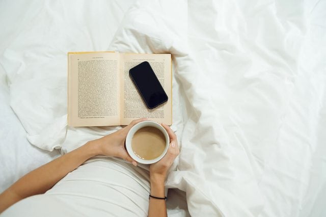 Female hands with a cup of coffee, phone and old book in white bed. Top view