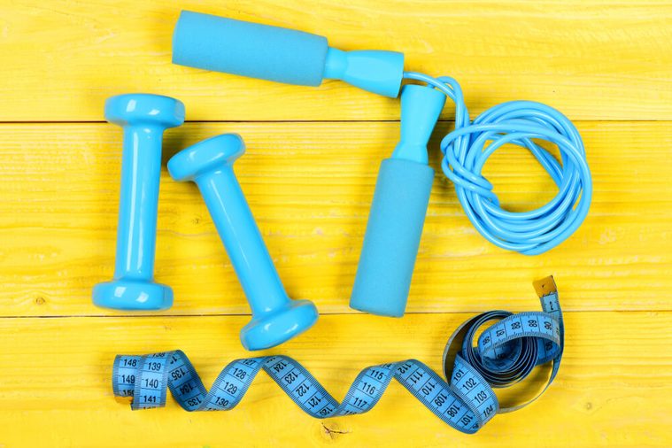Dumbbells, measuring tape and jump rope, top view. Sports equipment on wooden yellow background. Barbells and centimeter near tools for weight loss. Workout and active lifestyle concept.