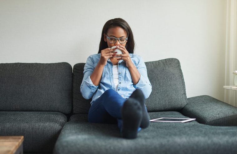 Young African woman sitting on her living sofa at home drinking a cup of coffee and relaxing