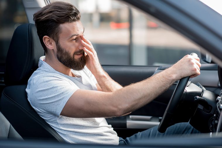 A bearded man driving and rubbing his eyes