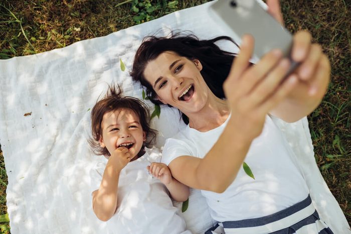 Happy brunette young smiling woman taking self-portrait to beautiful little girl daughter lying on white plaid on grass. Young family taking selfie outdoor. Motherhood, parenting and childhood concept