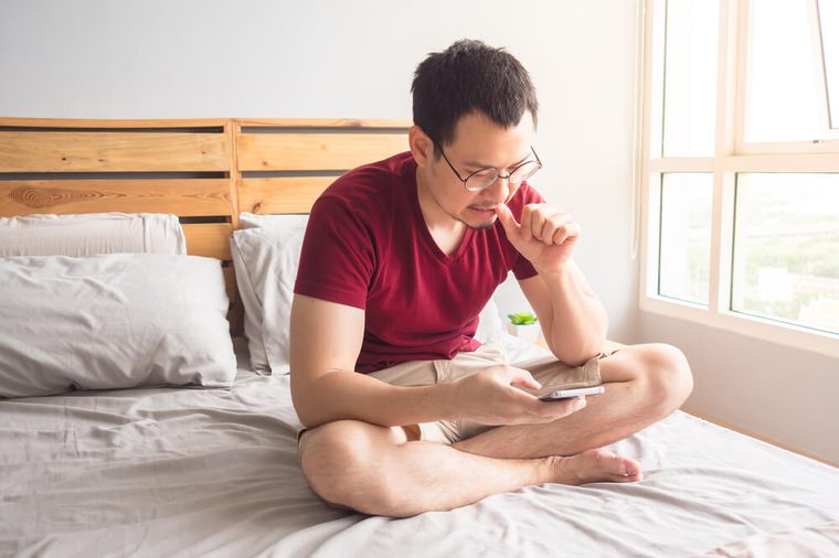 Lonely non-social Asian man with his smartphone in his bedroom apartment. Man bites nails.
