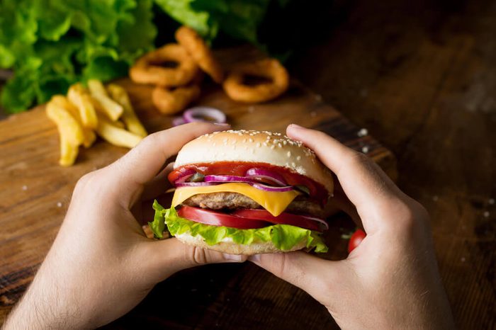 Cheeseburger. Man hands holding burger with cheese, red onion, tomatoes, lettuce green salad and pickles. Closeup view, selective focus