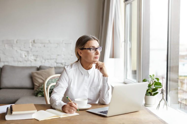 Attractive blonde mature female writer sitting at desk at home placing chin on her hands and looking away with thoughtful or unhappy expression while experiencing writer's block and creative slowdown