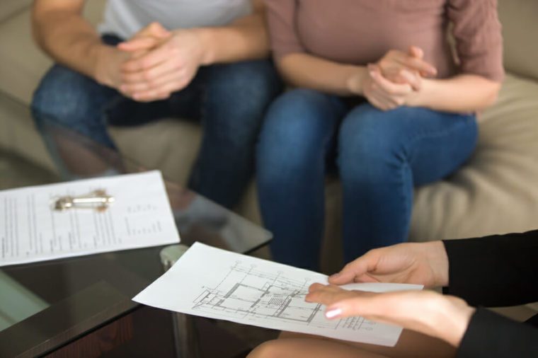 Close up view of female realtor showing to young couple of buyers flat plan on the sheet of paper sitting on sofa indoors, discussing buying apartment, considering flat purchase, interior layout 