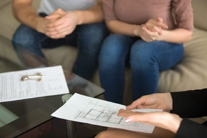Close up view of female realtor showing to young couple of buyers flat plan on the sheet of paper sitting on sofa indoors, discussing buying apartment, considering flat purchase, interior layout 