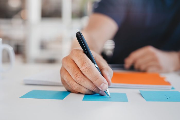 Close up image of business man writing on an adhesive note at table in office, focus on hand and pen.