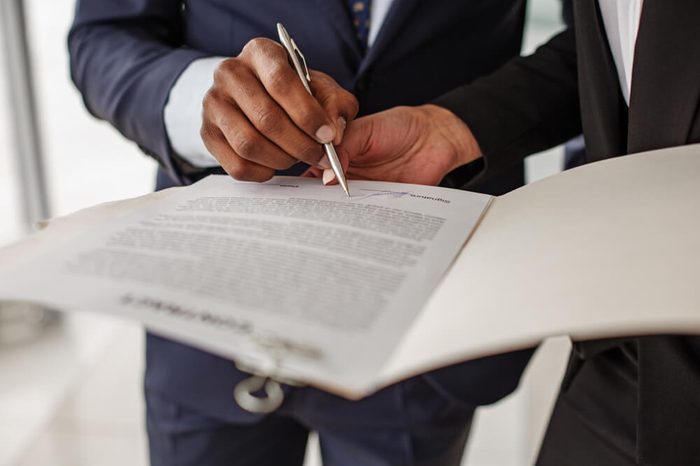 Sign it here. Woman is standing in office and holding folder with contract while man in suit is signing it. Close up of hand with pan. Selective focus