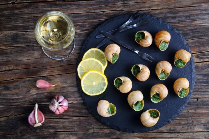 gourmet dish - Escargots de Bourgogne or Snails with herbs, butter, garlic on black slate plate with forks, ingredients and wine glass with dry white wine on wooden table, horizontal view from above
