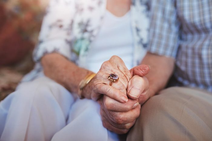 Cropped shot of elderly couple holding hands while sitting together at home. Focus on hands.
