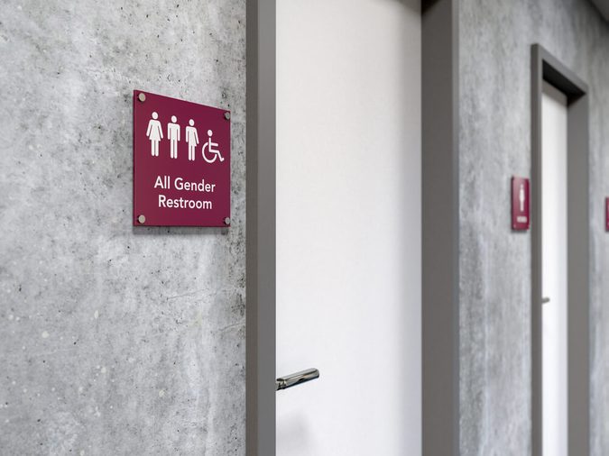 All-gender restroom signage next to a restroom door showing icons of man, woman, transgender and wheelchair user