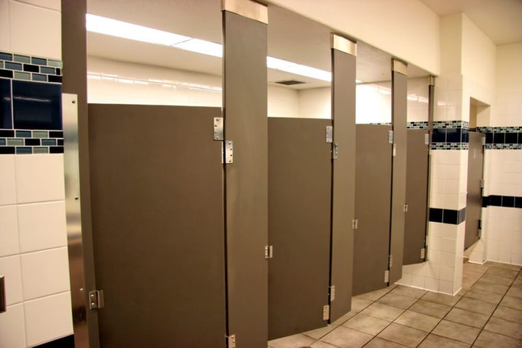 Public bathroom stalls. Four open brown metal stall doors. Fluorescent lighting in background. Beige tile floor and walls. Nobody. Horizontal.