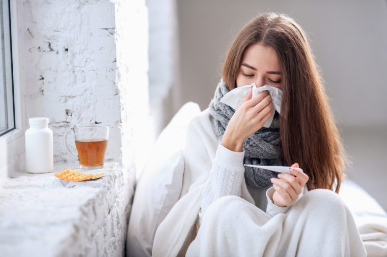 Cold and flu. Sick woman caught cold, feeling illness and sneezing in paper wipe. Closeup of beautiful unhealthy girl covered in blanket wiping nose and looking at thermometer. Healthcare concept.