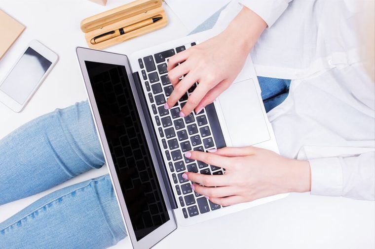 Woman in jeans sitting on white surface with smartphone, supplies and typing laptop keyboard placed on her lap. Mock up