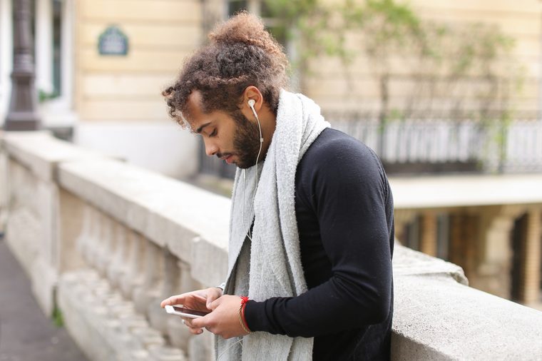 Mulatto tourist raving with white in ear headphones and smartphone near Eiffel tower. Jocund guy visiting Paris and enjoying with popular songs. Concept of using modern gadgets for entertainment and