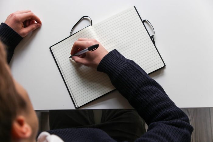 Student writes notes into notebook on white desk with black pen, top view