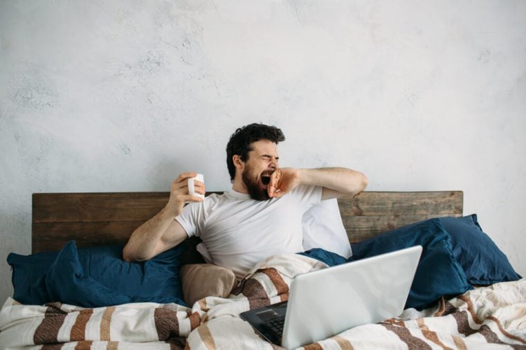 Handsome man lying in bed and warm blanket and stretching and yawning. Sleepy guy has laptop on the top and holding cup with hot tea or coffee in right hand