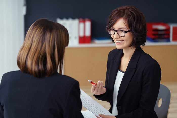 Cheerful Female Manager Talking to her Subordinate at her Table Inside the Office.